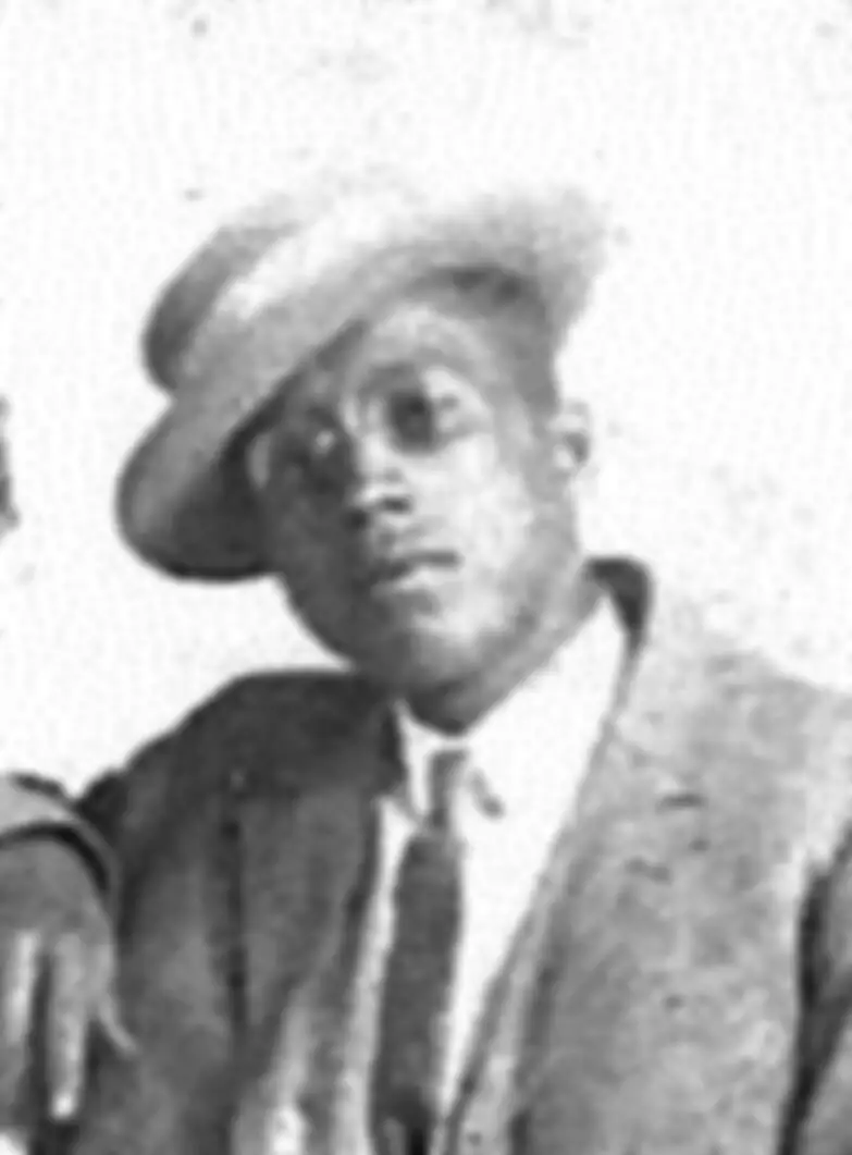 Black and white photo of a Black man in a suit and hat. His head is tilted to one side and his right elbow is propped up on something, right hand hanging down.