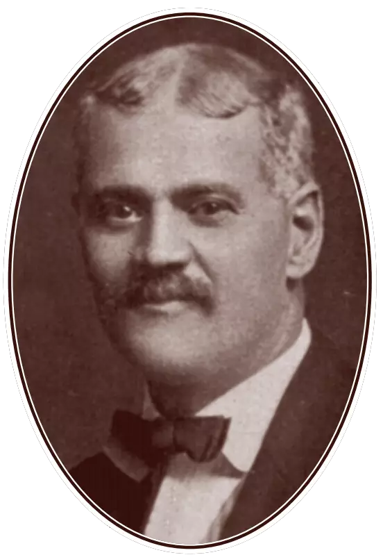 Picture of a man, shoulders up, wearing a suit and bow tie, with a mustache, looking directly into the camera.