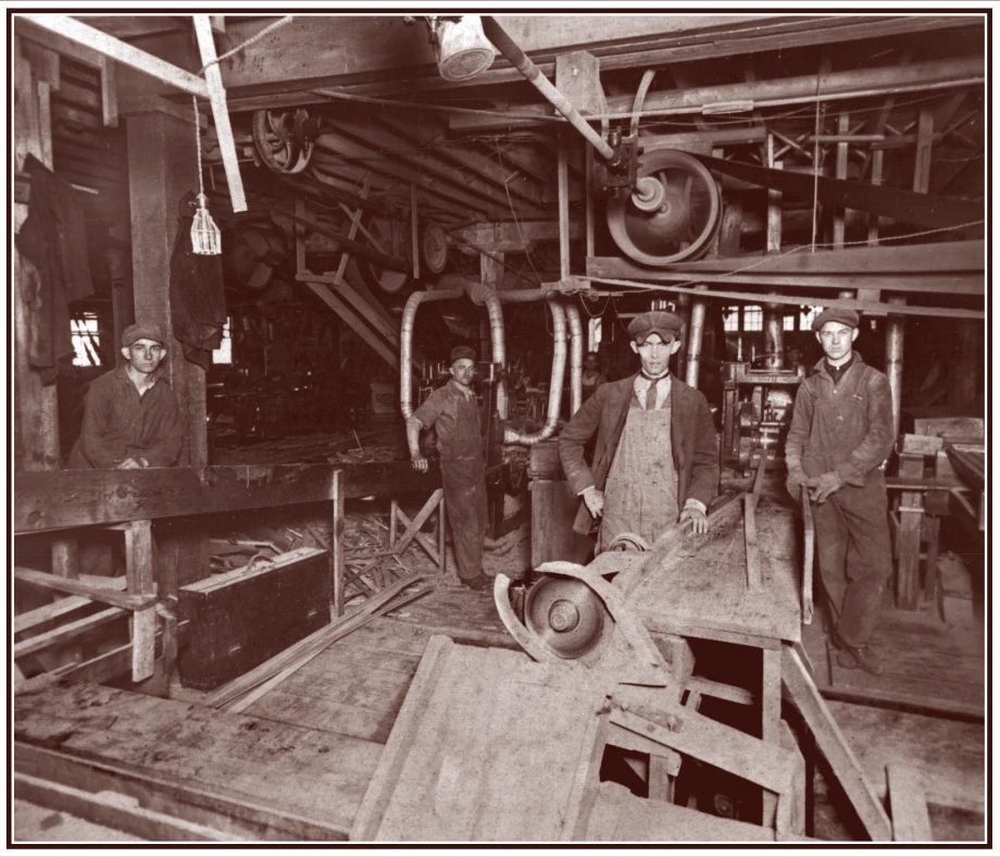 four men stand inside a wooden mill. Machinery is all around them, including on the ceiling. it looks dark.