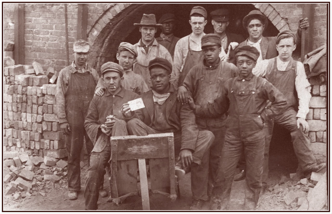 a group of young men and boys pose for a photograph under a brick archway. Bricks are stacked all around them.