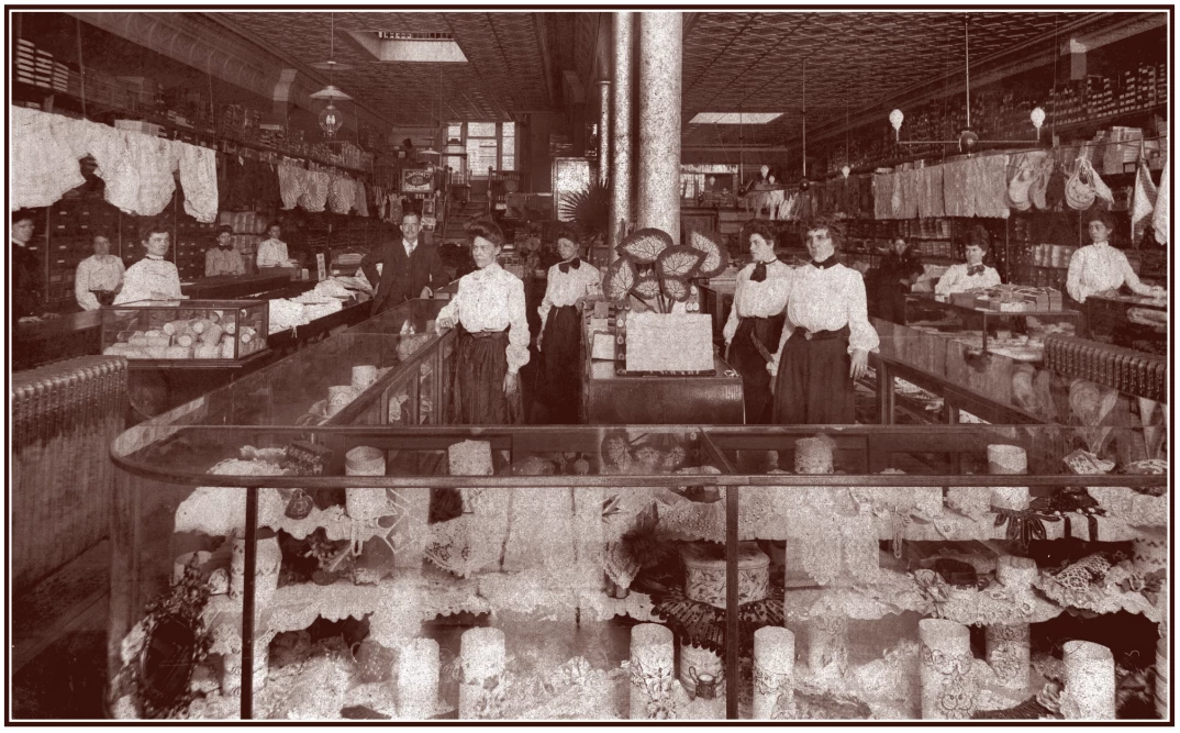 Portrait of women working at a clothing store. They are wearing white shirts and long dark dresses. They are surrounded by glass display cases and stacks of fabric on the back walls.