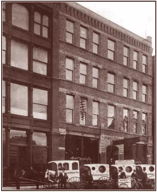 A four-story brick building with horse drawn white carriages in front. the carriage have large wheels and are white with Model Laundry Co. printed on the side. Men stand next to each carriage in suits and hats.