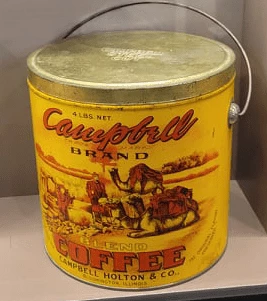 A color photograph of an antique coffee tin sitting on a pink table in front of a pink wall. The coffee tin is yellow with Campbell brand label with image of people and camels at a countryside.