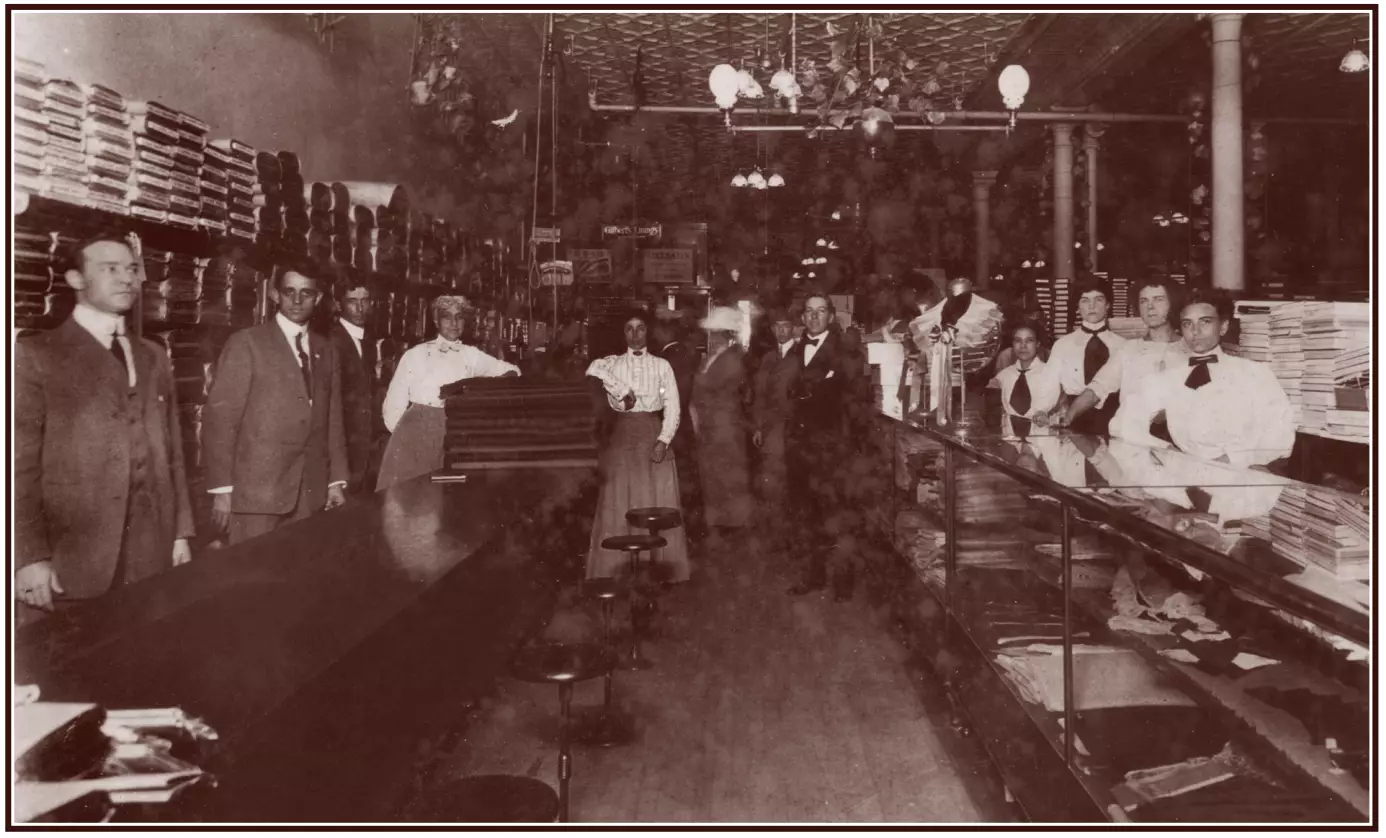 Portrait of men and women at work in a clothing store. One man is in a tuxedo, while other men wear three-piece suits. The women wear long dresses that reach the floor. Fabrics are stacked behind them, and clothing is displayed in glass cases.