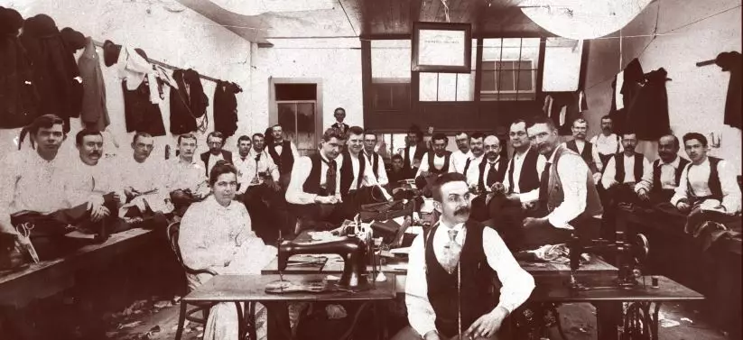 Black and white portrait of tailor shop employees. There is one woman and 27 men, all of whom are wearing vests, white shirts, and ties. The men are lined up along the walls, and there are sewing machines in the middle, where the woman is seated. The room is bright, and coats hang along the walls above where the men are seated.
