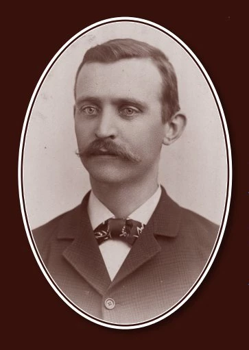Portrait of a young light-skinned man with short hair combed back, thick handlebar mustache, and prominent ears. He is wearing a dark suit with white shirt and bow tie.