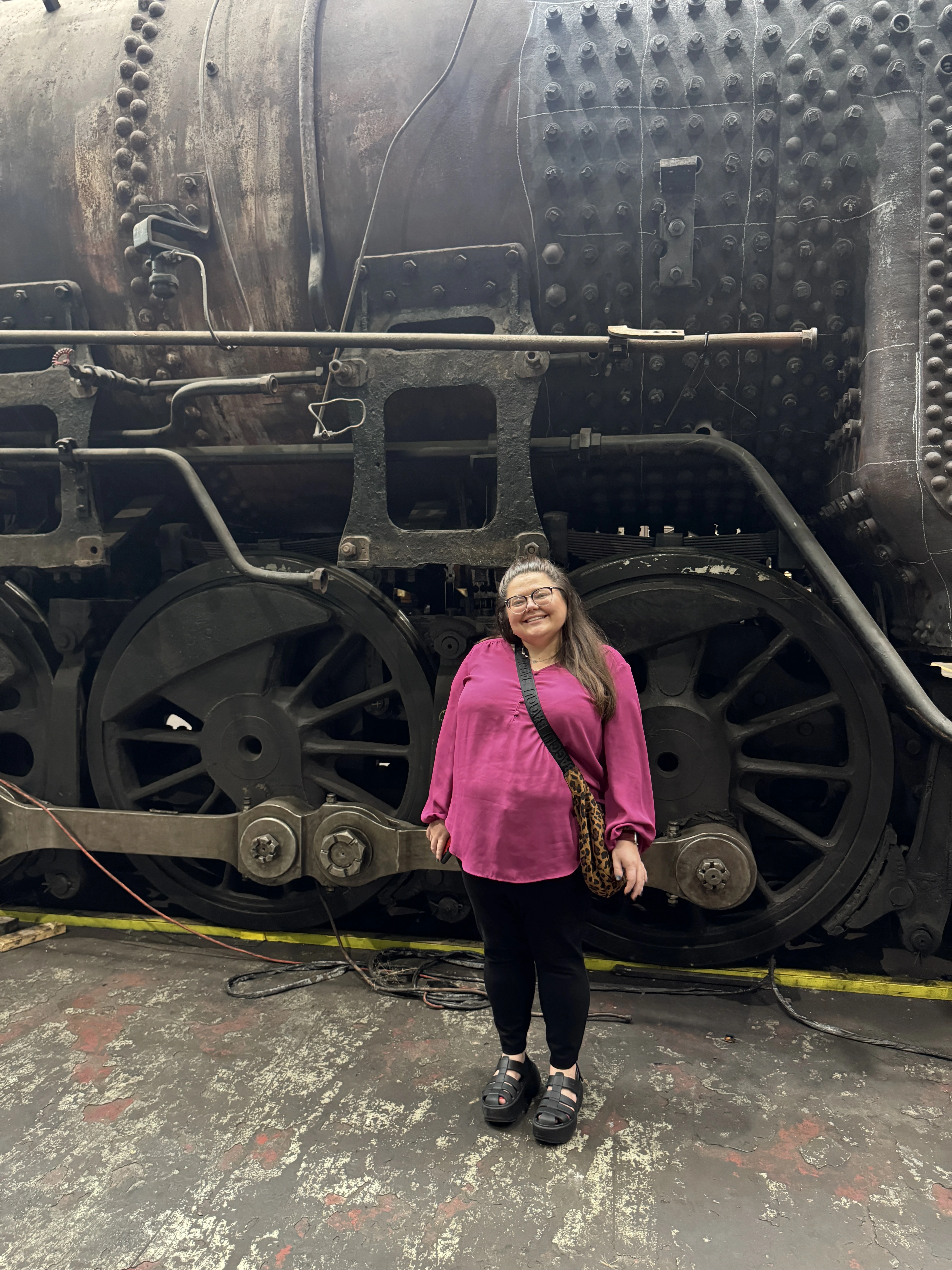 Maria stands in front of a steam engine wearing a pink shirt. She's just as tall as one of the wheels.