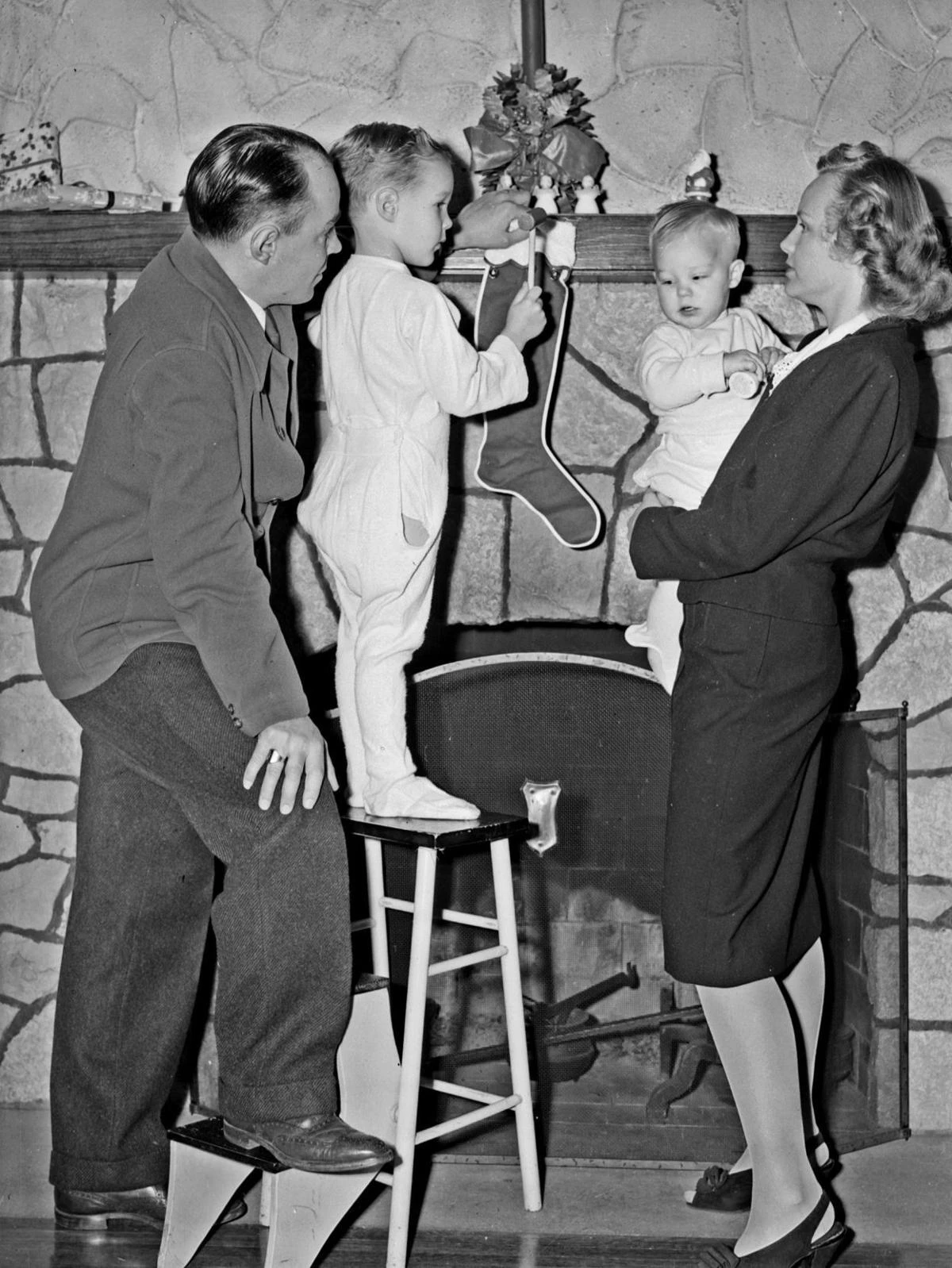 a family stands in front of a fireplace, looking at stockings hanging