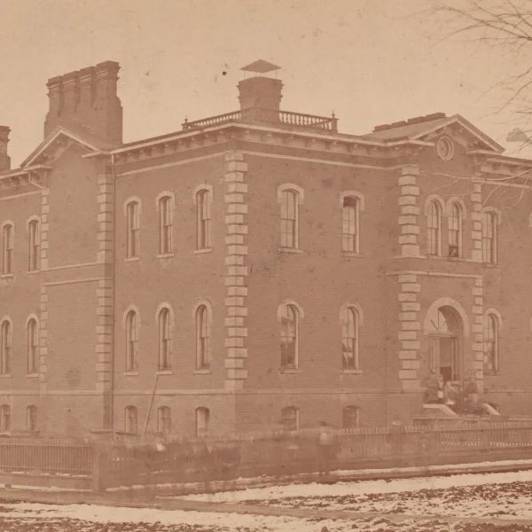 A yellowed image shows a large three-story school made of brick. it has multiple chimneys, and a fence around the perimeter of the grounds. Blurred human figures can be seen on the sidewalk, they are moving and come across as translucent in this image. A few branches of a tree can be see on the right, they are completely bare. A small amount of snow appears to be on the ground.