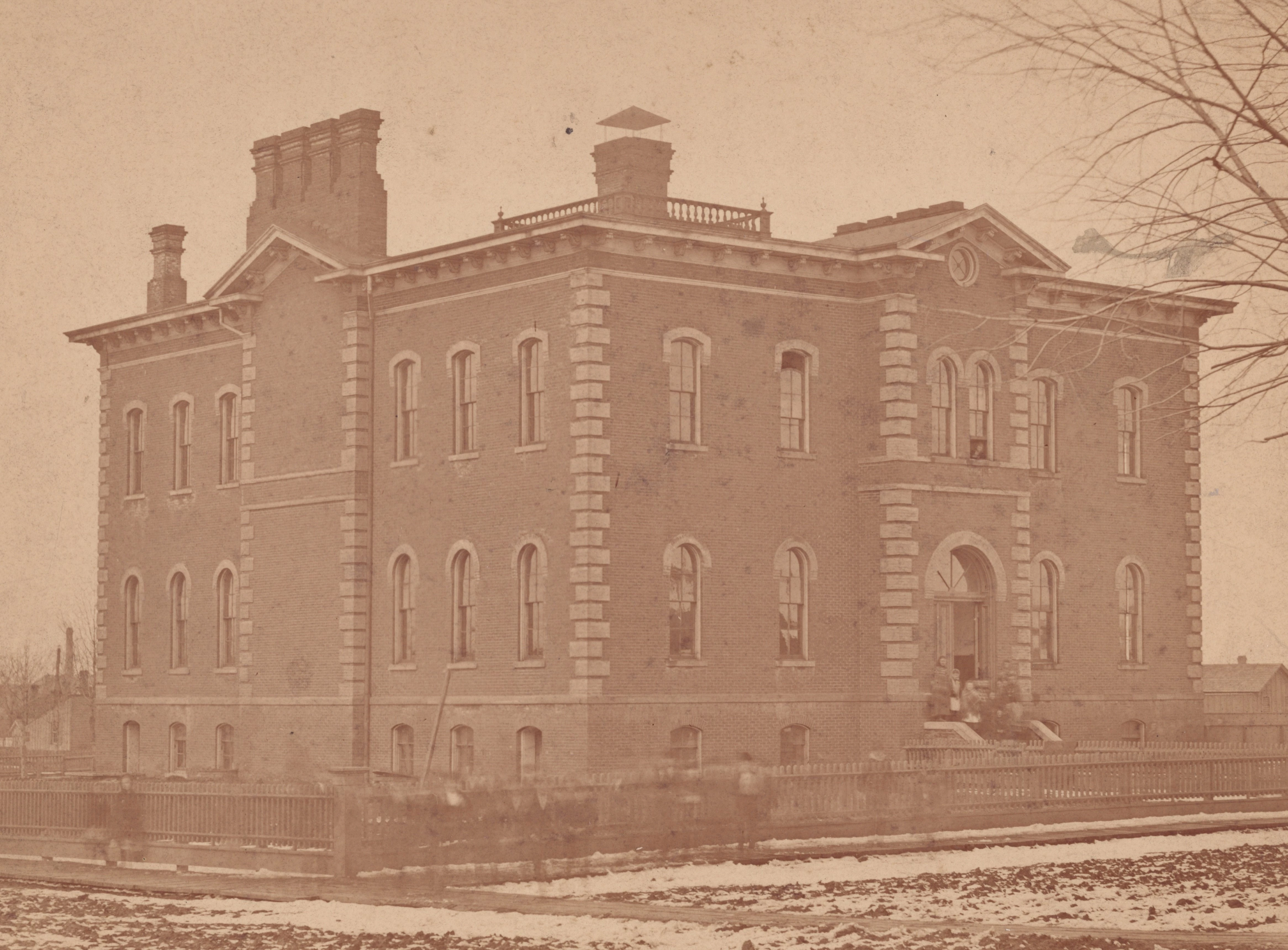 A yellowed image shows a large three-story school made of brick. it has multiple chimneys, and a fence around the perimeter of the grounds. Blurred human figures can be seen on the sidewalk, they are moving and come across as translucent in this image. A few branches of a tree can be see on the right, they are completely bare. A small amount of snow appears to be on the ground.