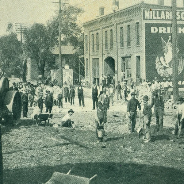 dozens of people are in this image, all standing looking towards the camera. Dirt is in the foreground, as well as a large steamroller. In the background is a two-story brick building, homes, trees, and utility poles. Many men are holding buckets and shovels.