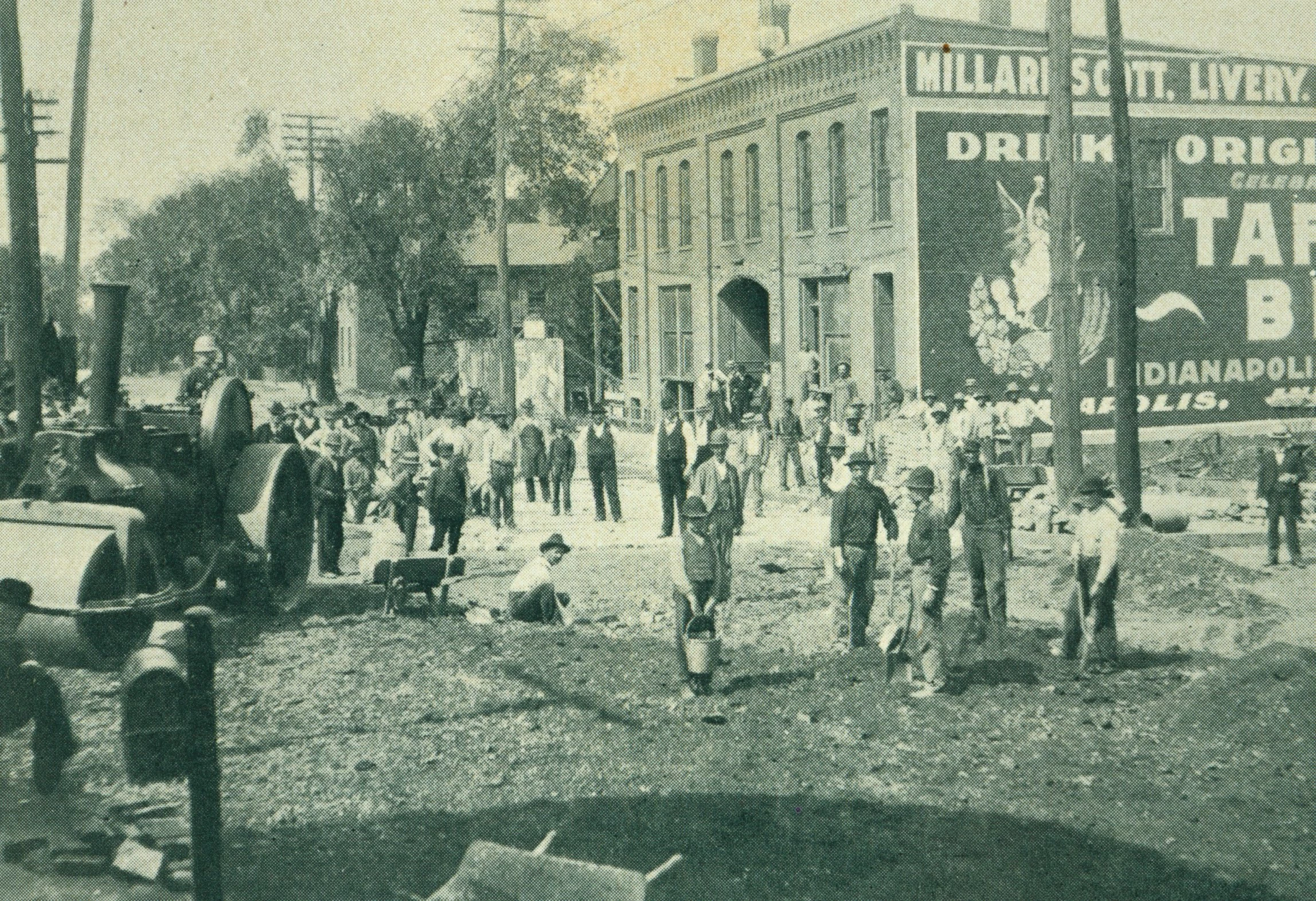 dozens of people are in this image, all standing looking towards the camera. Dirt is in the foreground, as well as a large steamroller. In the background is a two-story brick building, homes, trees, and utility poles. Many men are holding buckets and shovels.