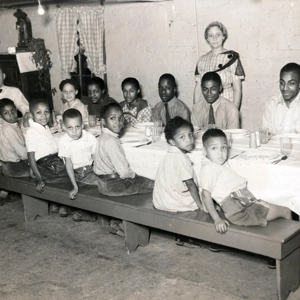 13 children and young adults sit on a long bench at a table. The table has a white table cloth and place settings for each person. One man is at the head of the table, wearing a bow tie. A woman stands behind the table in a dress. They are all looking at the camera, many of the children are smiling. All of the people in the photograph are African American. The walls appear to be lightly-painted brick.