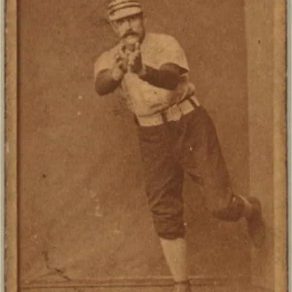 portrait of a man wearing a baseball uniform getting ready to catch a baseball. He has bare hands extended in front of him, and is leaning on his right leg, left leg bent in the air behind him.