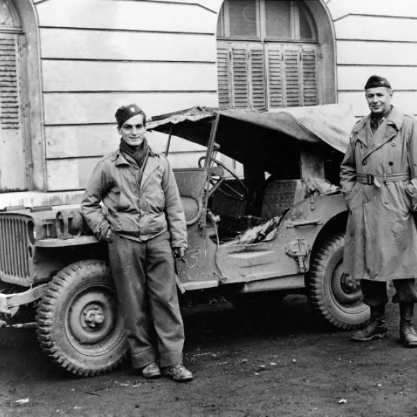 Two light-skinned men stand outside a military vehicle. They are parked on a dirt road with a masonry building with large arched windows behind them.