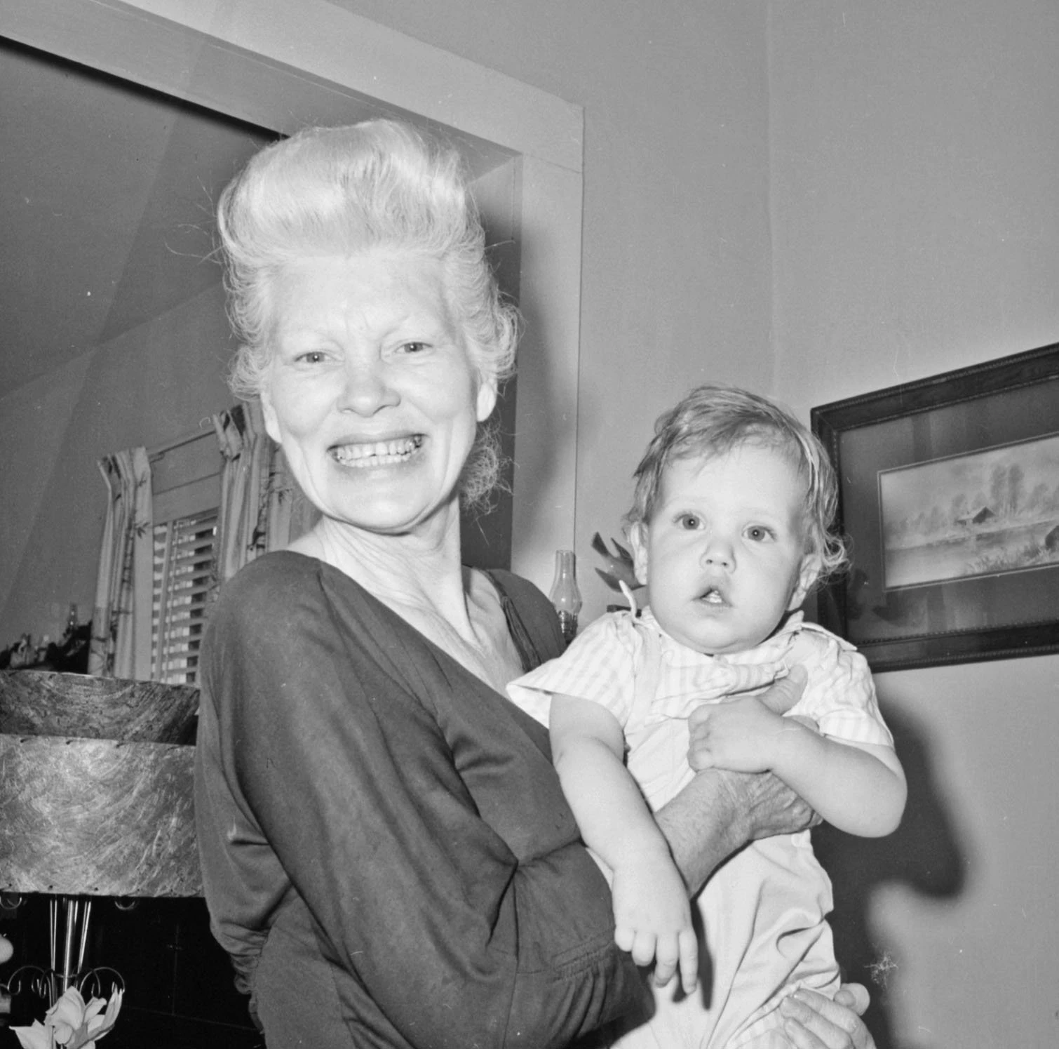 A light-skinned woman with blonde hair holds a young child. The woman has a big smile on her face and is looking straight at the camera. She is inside a home. The baby has a head full of hair and has his mouth open.