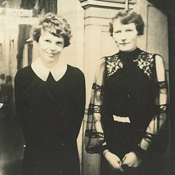 two light skinned women with short wavy hair stand next to each other looking at the camera. Amelia has a friendly closed-mouth smile and is wearing a long-sleeved black dress with large white collar. Next to her, Brenneman is wearing a dark dress with sheer sleeves.