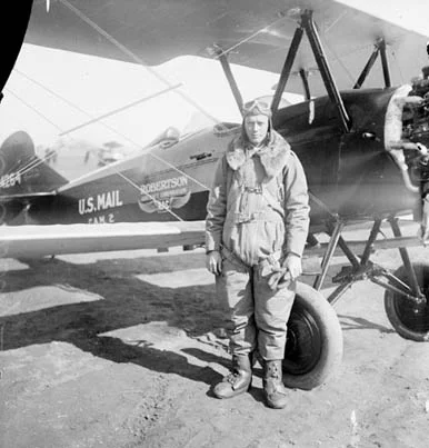 a man wearing an aviation suit stands outside a plane that says US MAIL on the side