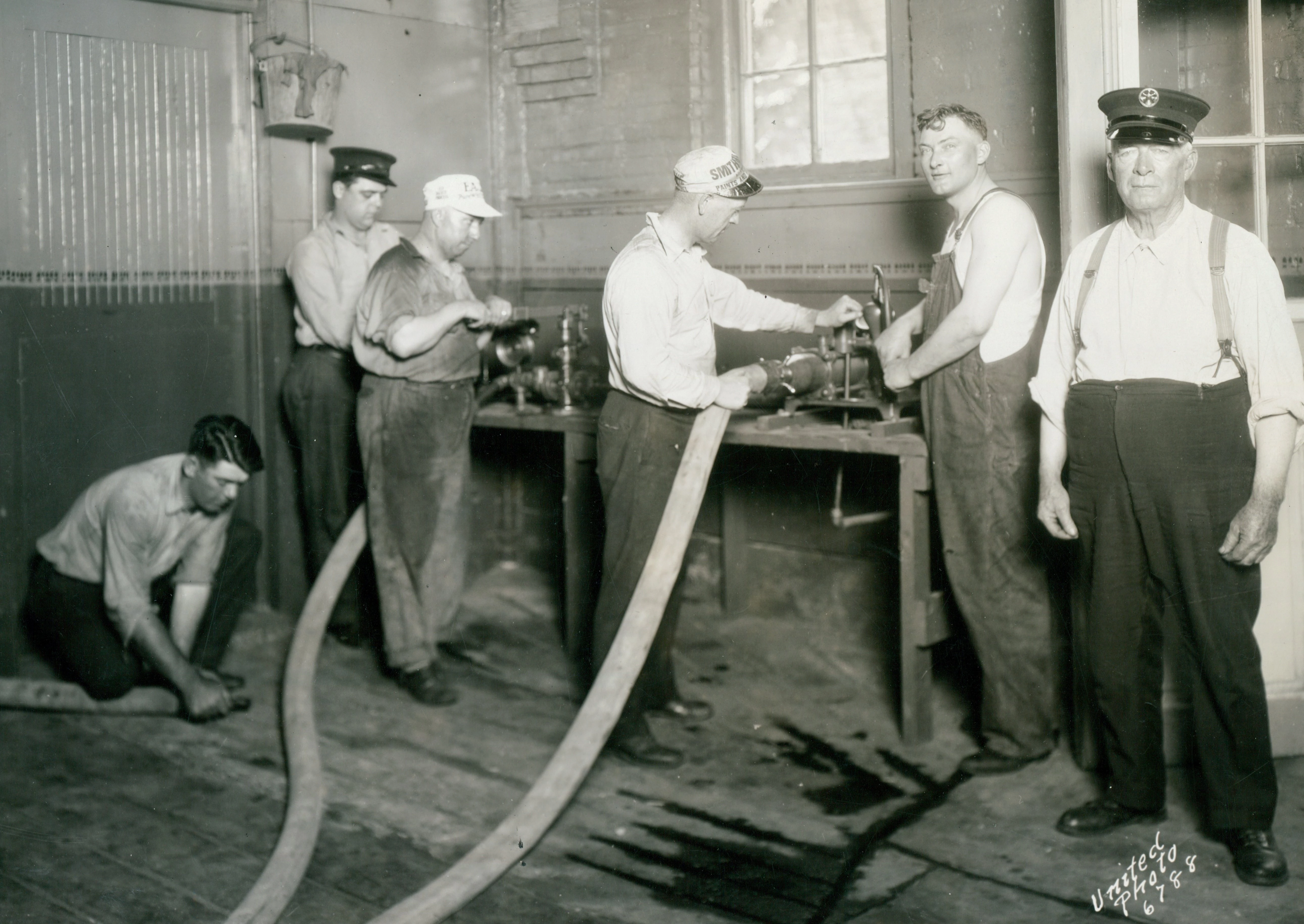 6 men are in this photograph, two standing at a contraption with hoses attached. The floor is wet.